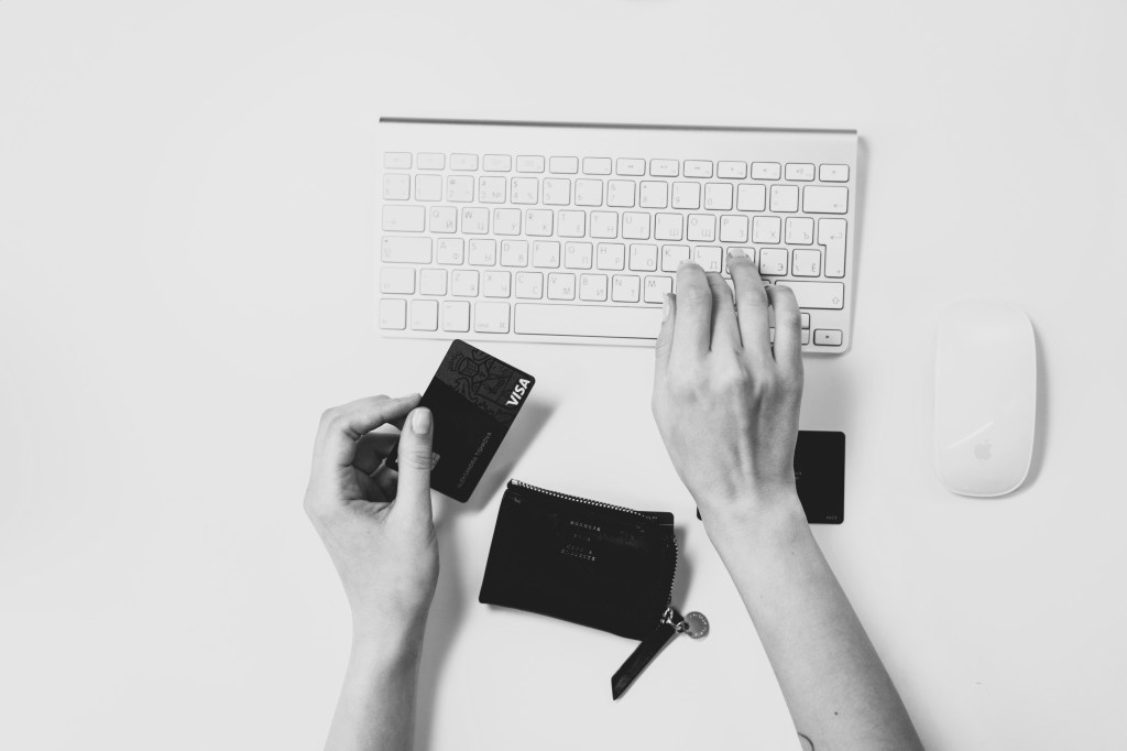 black and white image depicting the hands of a woman writing to a computer keyboard and holding a credit card